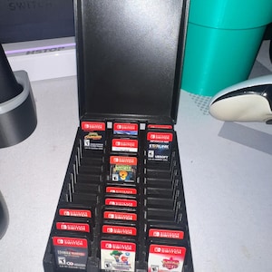 Może przedstawiać: Czarny, prostokątny pojemnik do przechowywania kartridży do gier, otwarty, aby odsłonić wiele kartridży do gier Nintendo Switch. Obudowa wykonana jest z matowego czarnego materiału i ma pokrywę na zawiasach. Kartridże są czerwone i czarne.