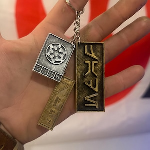 Könnte beinhalten: Ein silberner Schlüsselanhänger mit drei rechteckigen Anhängern. Ein Anhänger zeigt ein detailliertes Emblem, ein anderer stilisierte Schrift und der dritte eine Reihe von Symbolen. Der Schlüsselanhänger wird vor einer Flagge mit rotem und weißem Design gehalten.