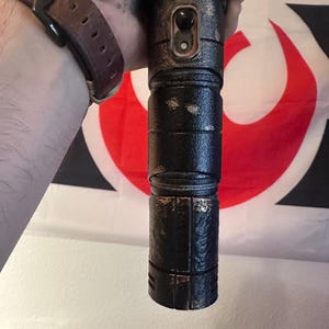 Puede incluir: Un sable de luz negro y bronce con un acabado texturizado. El sable de luz tiene un diseño cilíndrico con un botón y una serie de crestas. El fondo incluye una bandera roja, blanca y negra.