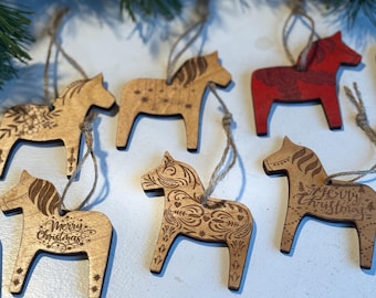 Puede incluir: Adornos de caballo de madera con intrincados diseños grabados y colgadores de cuerda. Algunos son de madera natural, otros están pintados de rojo. Un adorno tiene grabado "Merry Christmas". Decoración navideña.