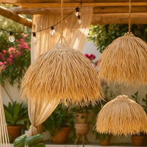 Puede incluir: Tres l&aacute;mparas colgantes con pantallas de paja natural cuelgan de una estructura de madera. Las pantallas tienen un borde con flecos y las luces est&aacute;n suspendidas por cuerdas trenzadas. Hay guirnaldas de luces sobre las pantallas.
