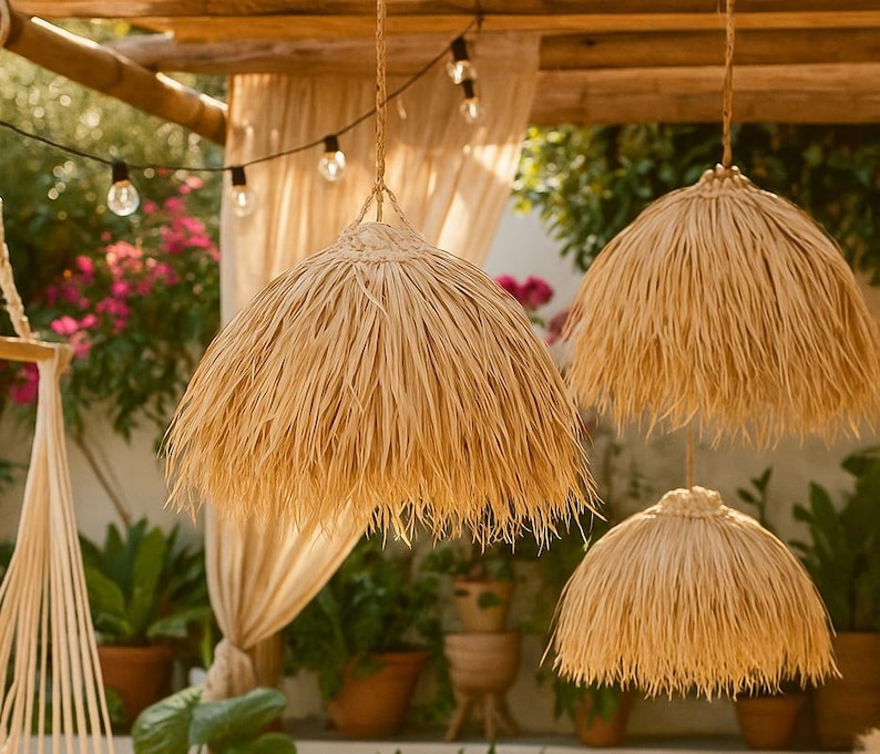 Puede incluir: Tres l&aacute;mparas colgantes con pantallas de paja natural cuelgan de una estructura de madera. Las pantallas tienen un borde con flecos y las luces est&aacute;n suspendidas por cuerdas trenzadas. Hay guirnaldas de luces sobre las pantallas.