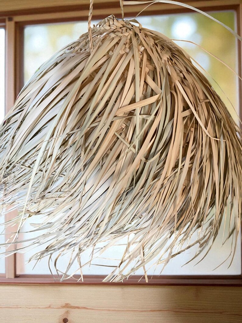 Puede incluir: Una l&aacute;mpara colgante natural y tejida. La pantalla est&aacute; hecha de hojas de palma secas de color marr&oacute;n claro, creando un efecto texturizado y en capas. La l&aacute;mpara est&aacute; suspendida de una parte superior tejida, proyectando un brillo c&aacute;lido y acogedor.