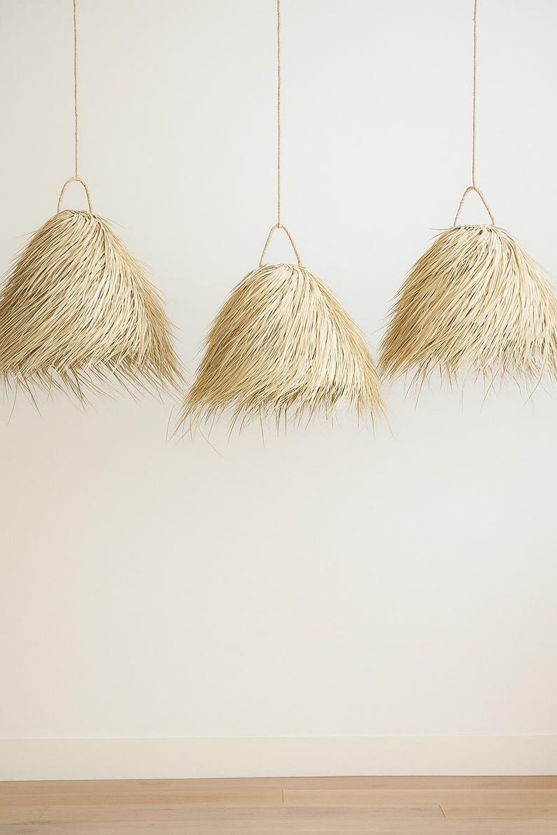 Puede incluir: Tres l&aacute;mparas colgantes con pantallas de paja natural suspendidas de finos cordones. Las pantallas tienen forma c&oacute;nica con un borde con flecos, y cuelgan contra una pared neutra. Las luces son de un color c&aacute;lido y neutro.