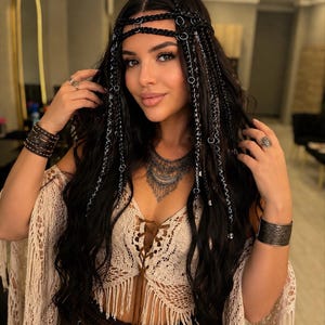 May include: A black hair accessory featuring braided strands, silver-toned rings, and metallic accents worn across the forehead. The piece includes multiple dangling chains and braided sections that integrate into long, dark, wavy hair.