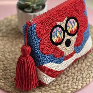 May include: A rectangular punch needle pouch featuring a textured design of a character with red hair and round glasses with colourful zigzag patterns. The pouch has a red zip closure with a large red yarn tassel attached to the side zip pull.