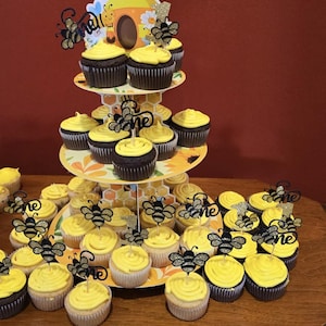 May include: A tiered cupcake stand with yellow frosted cupcakes, some chocolate and some vanilla. Each cupcake is decorated with a black and gold glitter bee and the word "one". The stand is decorated with a bee and honeycomb theme.