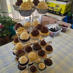 May include: A tiered cupcake stand displays an assortment of cupcakes with chocolate and vanilla frosting. The cupcakes are in silver foil liners and topped with "Happy Anniversary" toppers. The stand is on a table with a light blue and white checkered tablecloth.