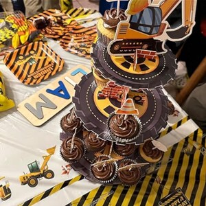 May include: A construction-themed cupcake stand with chocolate frosted cupcakes. The stand has tire-shaped tiers and construction vehicle toppers. Construction-themed plates and a wooden sign that says "AM" are also visible.