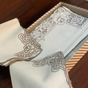 Puede incluir: Un juego de servilletas de lino blanquecino con un diseño floral en una caja decorativa. Las servilletas tienen un borde floral marrón detallado. La caja es beige con rayas marrones.