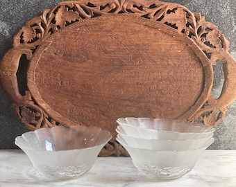 May include: A carved wooden serving tray with intricate floral designs and handles, paired with four frosted glass bowls with scalloped edges. The bowls are stacked on a white marble surface.