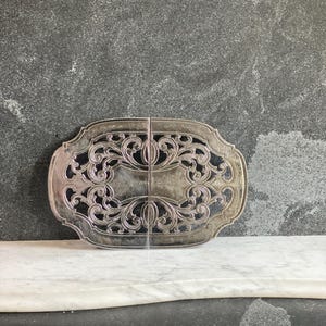 May include: Silver-toned, ornate serving tray with a symmetrical, openwork design. The tray is oval-shaped with a decorative border and a central pattern of swirling lines and curves. The tray is displayed on a white marble surface against a gray background.