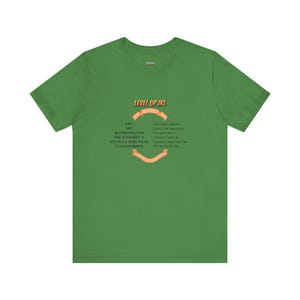 May include: Green t-shirt with the text "LEVEL UP IRL" in orange. Below the text are the words "HP, MP, STRENGTH, DEXTERITY, INTELLIGENCE, CHARISMA" and a list of phrases. The shirt is made of a soft material.