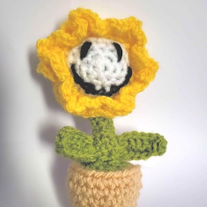 May include: A crocheted flower with a smiling face. The flower has yellow petals, a white face with a black smile, and a green stem with leaves. It sits in a light orange pot. The craftwork is detailed and whimsical.