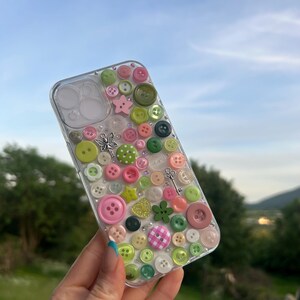 May include: A clear phone case decorated with a variety of pink and green buttons in different sizes and shapes. The design includes small silver charms shaped like a fairy and a key, along with scattered clear rhinestones across the surface.