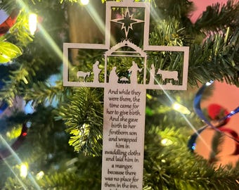 May include: Wooden Christmas ornament in the shape of a cross, featuring a star and Nativity scene cut-outs. The ornament includes the text "And while they were there...Luke 2, 6,7". It is hanging on a Christmas tree with green needles and colorful decorations.