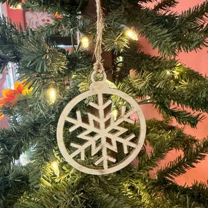 Pode incluir: Um enfeite de Natal de madeira em forma de floco de neve, pendurado por um fio de barbante. O enfeite está em frente a uma árvore de Natal verde com pequenas luzes brancas.