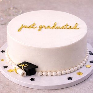 Op de afbeelding: Een wit geglazuurde taart met de gouden tekst "just graduated". De taart is versierd met een afstudeerhoed, een diploma en stervormige confetti. Een parelrand omlijnt de basis van de taart.