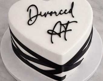May include: A heart-shaped cake with white icing and black ribbon detailing. The cake is inscribed with the words "Divorced AF" in black script. The cake is presented on a white cake board.