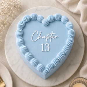 May include: A heart-shaped cake, iced in light blue with piped swirls around the edge. The cake is inscribed with "Chapter 13" in white script. It rests on a white marble plate, a celebratory dessert.