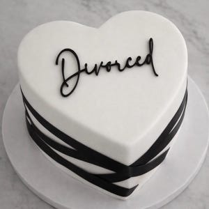May include: A heart-shaped cake with white icing and black ribbon detailing. The word "Divorced" is written in black cursive on the cake's surface. The cake is presented on a white cake stand.