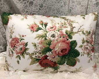 May include: Rectangular pillow with a white background and a floral pattern. The pattern features large pink and red roses, white flowers, and green leaves. The pillow is likely made of fabric and is a decorative item.