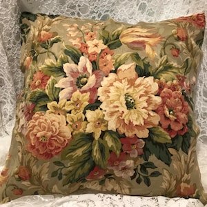 May include: Square decorative pillow with a floral pattern. The pillow features a beige background with a large arrangement of flowers in shades of peach, pink, yellow, and green. The flowers are detailed and realistic.