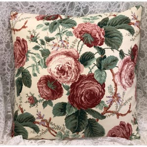 May include: Square decorative pillow with a cream background and a floral pattern. The pattern features large, detailed roses in shades of burgundy and pink, with green leaves and stems. The pillow is likely for home decor.