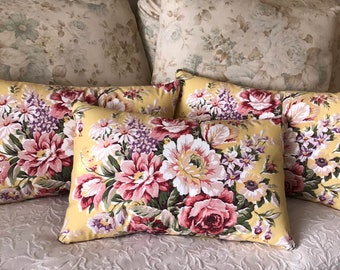 May include: Four yellow throw pillows with a floral pattern featuring pink, white, and purple flowers. The pillows are arranged on a bed with a floral patterned bedspread.