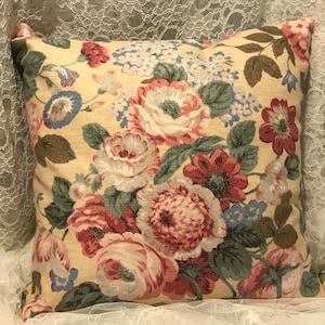 May include: Square decorative pillow with a floral pattern. The pillow features a cream-colored background with a large print of pink, red, and blue flowers and green leaves. The pillow is square-shaped.