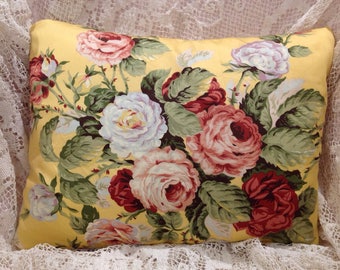 May include: A rectangular throw pillow featuring a vibrant yellow background with a dense, colorful floral pattern of blooming roses in shades of deep red, soft pink, and white, accompanied by detailed green leaves and foliage throughout the design.