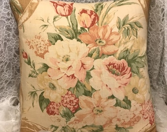 May include: A beige pillow with a floral pattern featuring pink, peach, and yellow flowers with green leaves. The pillow has a brown border.