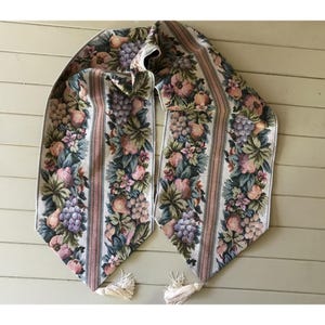 May include: A white table runner with a floral pattern featuring pink, green, and purple grapes, pears, and flowers. The runner has a pink and white striped background and white tassels on each end.