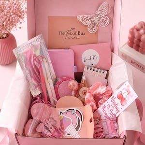 May include: A pink gift box filled with stationery and accessories, including a butterfly cutout, a notebook, a Drink Me sticker, a pink scrunchie, a bookmark with cherries and the text Reading is my therapy, and a unicorn ice cream sticker.