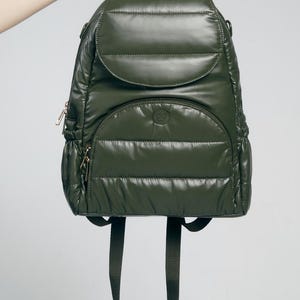 Peut inclure: Sac à dos vert olive avec un design matelassé. Le sac à dos a un rabat arrondi, une poche avant zippée et des bretelles réglables. Il comporte une poignée supérieure et des fermetures éclair dorées. Le sac est tenu par une main.