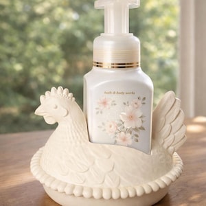 May include: A white foaming hand soap dispenser with a floral design, nestled in a cream-colored ceramic hen-shaped holder. The dispenser has a gold band and the text "bath & body works". The hen holder has a detailed design.