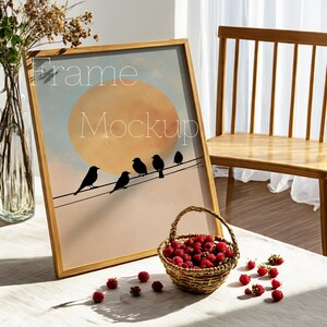 May include: A framed art print featuring a sunset scene with birds perched on a wire. The frame is made of light-colored wood. A small woven basket filled with red raspberries sits in front of the frame. The words "Frame Mockup" are printed on the art.