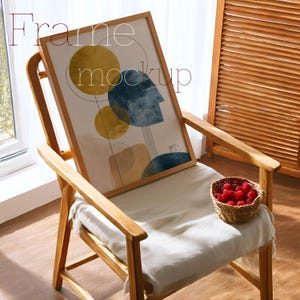 May include: A framed art print with abstract shapes in yellow, blue, and beige rests on a wooden chair. The artwork features a design with circles and a profile. The chair has a white cushion and a small basket of red berries.