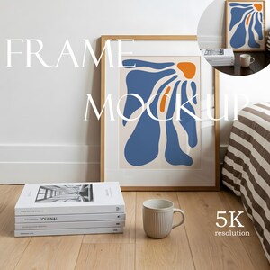 May include: A light wood picture frame containing an abstract print with blue and orange organic shapes on a cream background. The frame is displayed alongside a stack of four white books and a ribbed ceramic mug on a light wood floor.