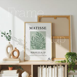 May include: A framed art print featuring a Matisse design with the text "MATISSE" and "BERGRUEN & CIE". The artwork has a light green botanical illustration on a white background. The frame is light brown and is displayed on a wooden shelf.