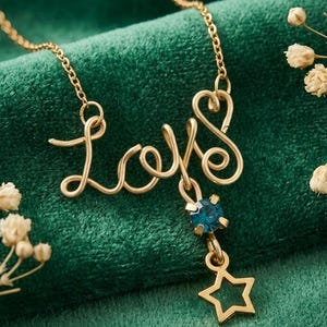 May include: A gold necklace spelling out "Love" in a cursive font, with a blue gemstone and a star pendant. Set against a dark green backdrop, the necklace is inscribed with "Shree Radhey creation" at the bottom.
