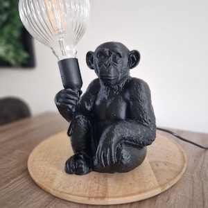 Puede incluir: Una lámpara negra con forma de chimpancé con una bombilla grande, transparente y acanalada. La lámpara está sobre una base redonda de madera. La lámpara está sobre una mesa de madera.