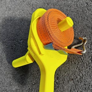 Può includere: Una cinghia a cricchetto giallo brillante con una cinghia arancione arrotolata all'interno. La cinghia ha un gancio di metallo all'estremità. Il meccanismo a cricchetto è visibile. L'articolo è su una superficie grigio scuro.