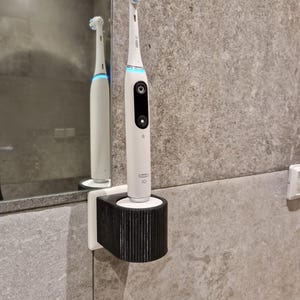 May include: A white electric toothbrush with a blue brush head is held in a black and white holder on a grey tiled wall. The toothbrush has a power button and a small display. The holder is rectangular with vertical lines.