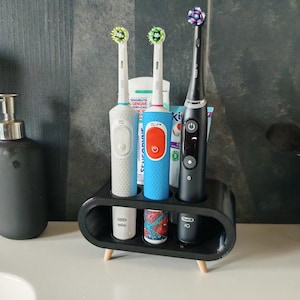 Puede incluir: Un soporte negro para cepillos de dientes con tres cepillos de dientes eléctricos y un tubo de pasta dental. El soporte tiene un diseño redondeado con patas de madera. Los cepillos de dientes son blancos, azules y negros con cabezales de diferentes colores.