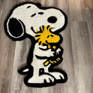 Peut inclure: Tapis Snoopy blanc et noir avec le personnage jaune de Woodstock étreignant Snoopy. Le tapis a une bordure noire et est posé sur un sol en bois. C'est un article de décoration intérieure sur le thème des dessins animés.