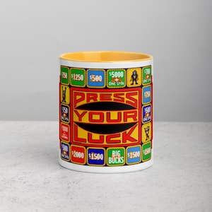 May include: A white ceramic mug with a yellow interior. The mug features the text "PRESS YOUR LUCK" in a red and yellow design, surrounded by boxes with dollar amounts and cartoon characters. The mug is a fun, novelty item.