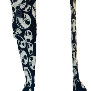 May include: A pair of black braces featuring a repeating white pattern of skull faces with stitched mouth details. The braces include a small rectangular fabric tab on one strap secured by a single black button, and metal hardware.