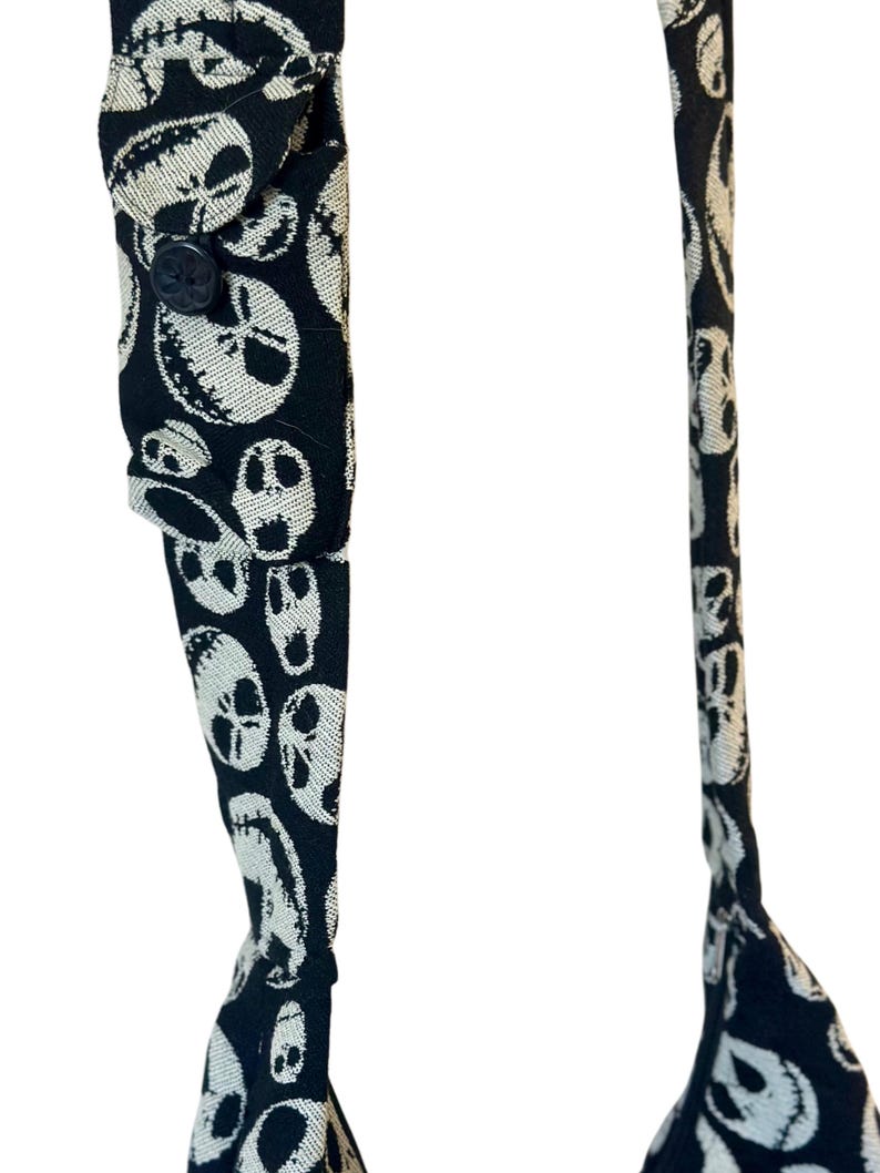 May include: A pair of black braces featuring a repeating white pattern of skull faces with stitched mouth details. The braces include a small rectangular fabric tab on one strap secured by a single black button, and metal hardware.