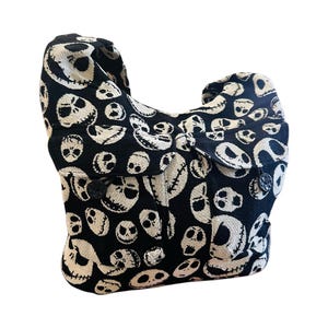 May include: A black and off-white hobo bag featuring an all-over woven pattern of various skull faces with stitched-style mouths. The bag has a single shoulder strap, a slouchy silhouette, and two front pockets secured with black flower-shaped buttons.